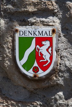 Blechschild mit Schriftzug "Denkmal" und Wappen Nordrhein-Westfalens, angeschraubt an einer Mauer