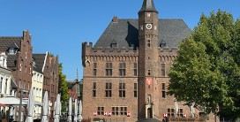 Backsteinbau mit mittig herausragendem Treppenturm, vorgelagert der Mrktplatz mit einem Baum