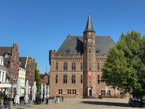 Backsteinbau mit mittig herausragendem Treppenturm, vorgelagert der Mrktplatz mit einem Baum